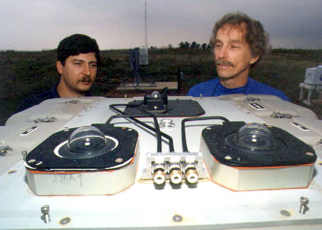 the heads of two mustachioed people peek up to look over white paneling with black tubing and instrumentation