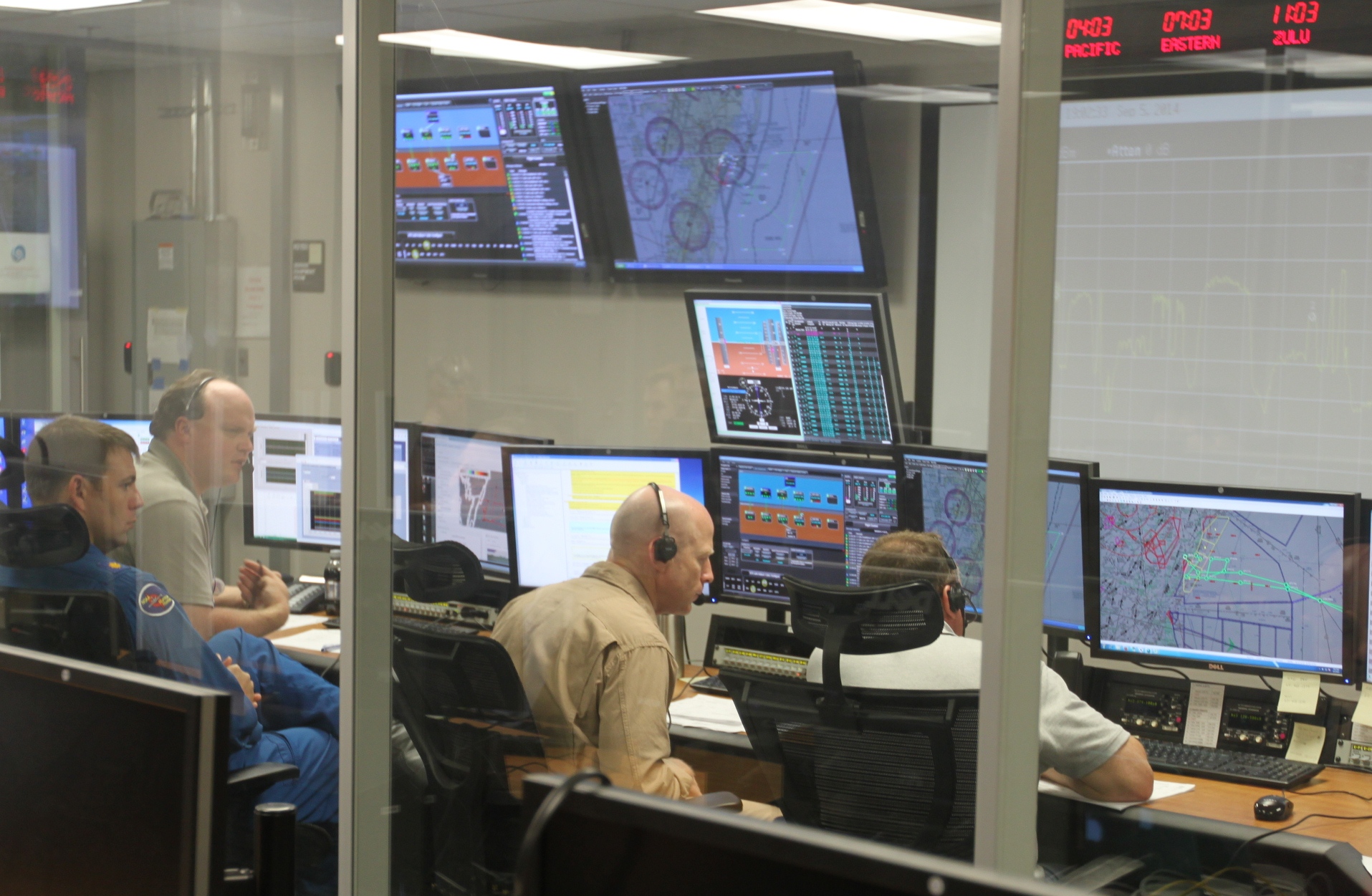 A view of a beige room full of computer monitors, manned by four men, is seen through a window. Beige window bars run top-to-bottom, outlining the center third of the photo. In the center photo are two of the men conversing, the one on the left is in a tan shirt and the man on the right is in a gray polo. In the left third, a man in a blue jumpsuit sits on the right next to another man in a tan shirt. Both of them are watching the men in the center third.