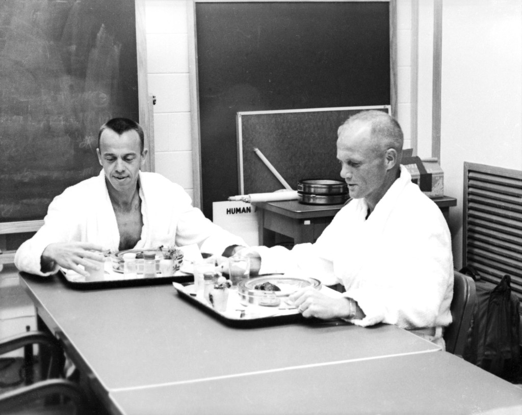 Alan Shepard and John Glenn, wearing white robes, are seated at a table with trays with their breakfasts