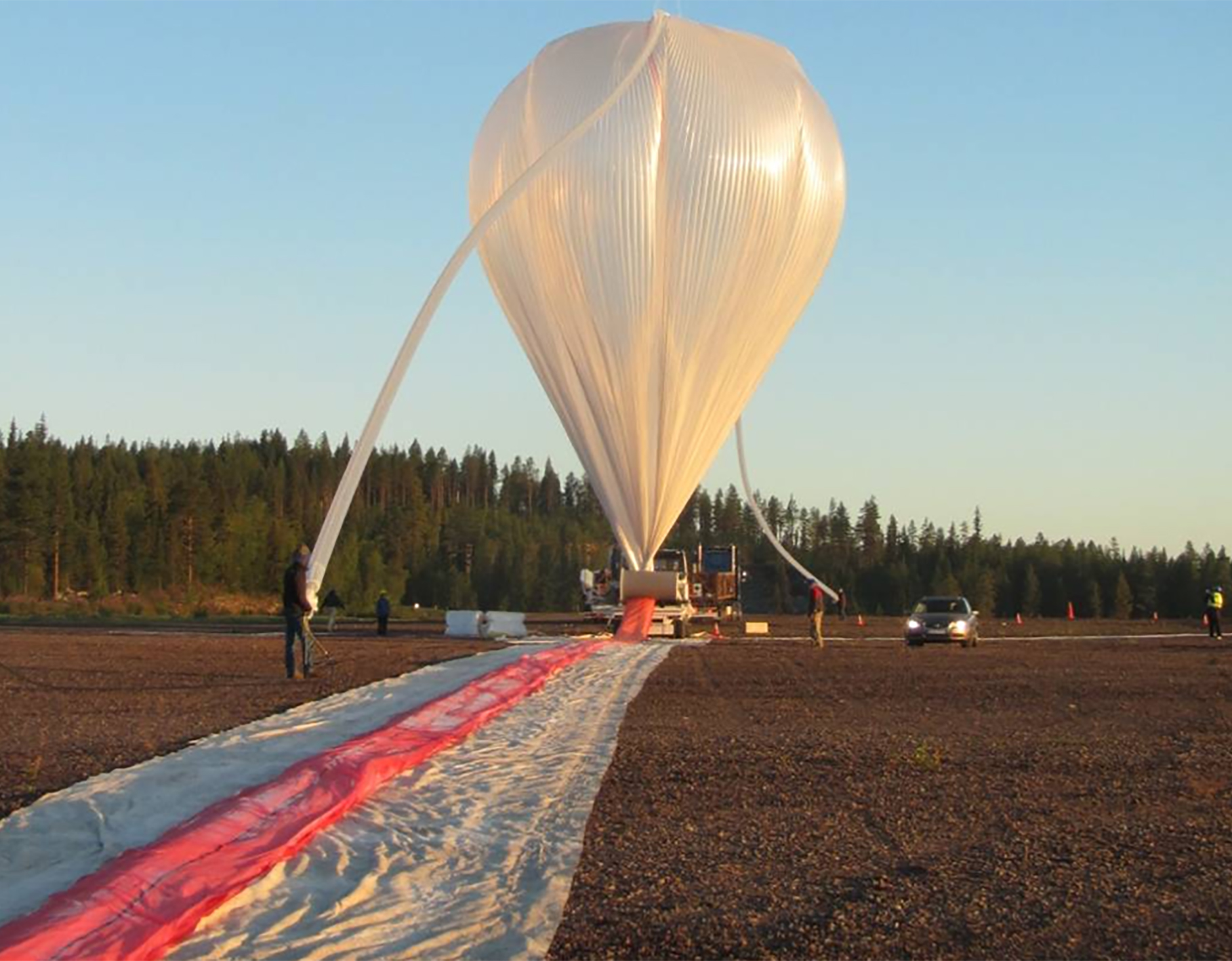 A gossamer white balloon fills the center of the image, with the brown dirt bottom third of the image covered in the unfilled area of the ballon. Above the balloon trail on the left are two people, one closer to the foreground, silhouetted against a curtain of pine trees that divides the pale blue dawn sky from the dirt. to the right of the inflated balloon is a small dark sedan with the headlights on and a man in a red jacket.