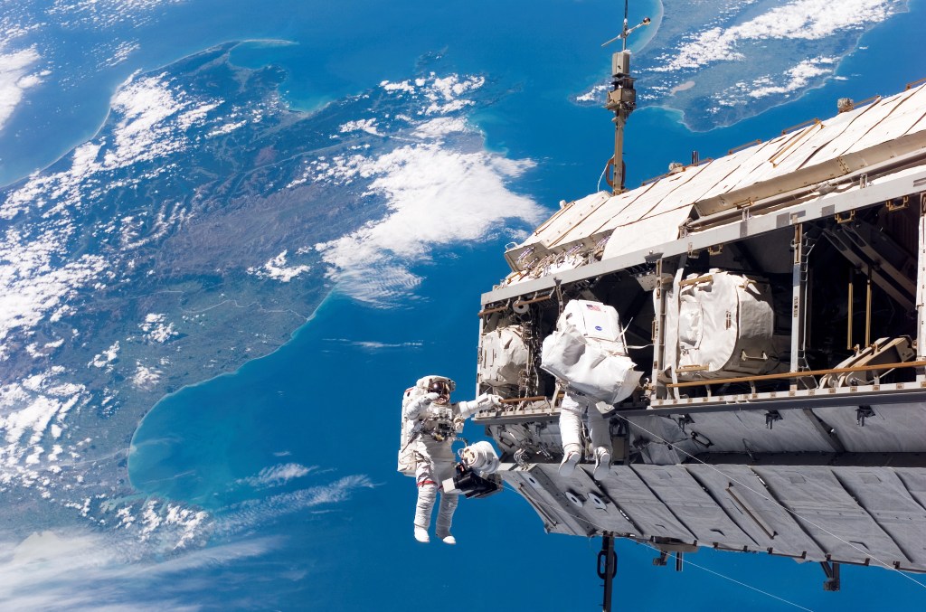 Astronauts working in space on a large piece of hardware with the Atlantic Ocean and Eastern United States visible in the background