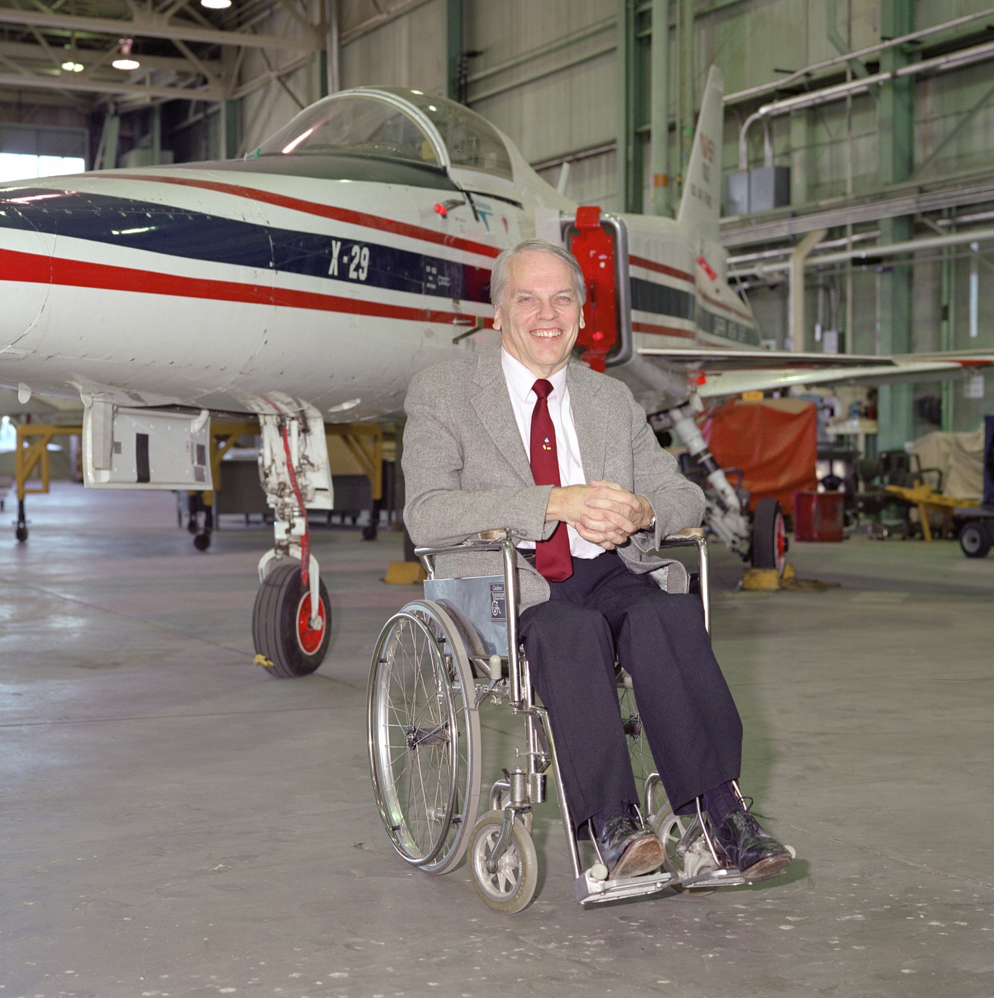 A man sits in a wheelchair with an experimental aircraft in the background.