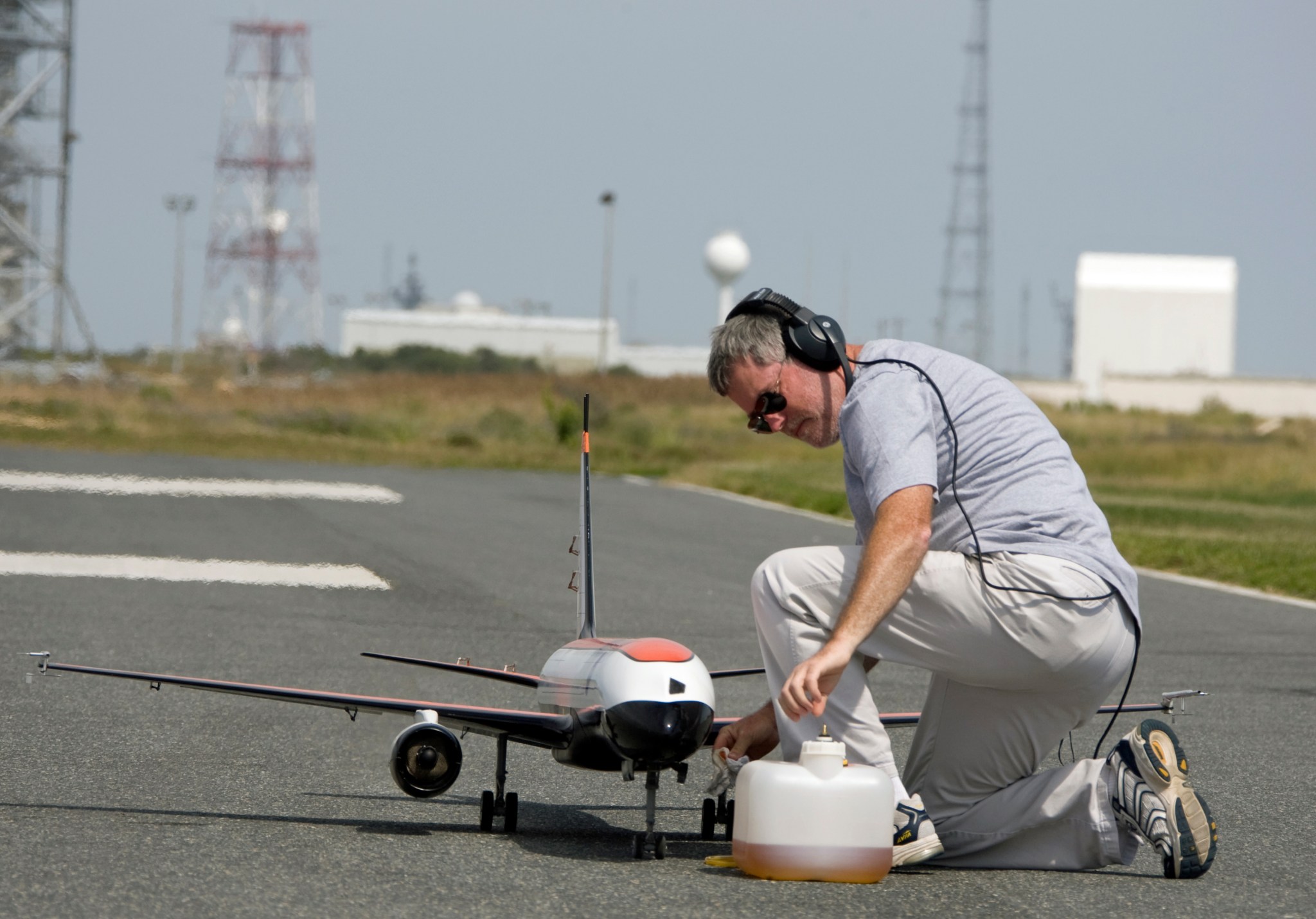 A man in a grey shirt, white pants, and black over-the-ear headphones kneels to the right of a remote control model plane on a runway at Wallops Flight facility. The white model plane has orange and black accents.