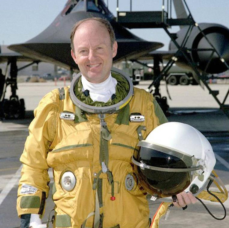 Man in space suit, holding helmet and posed smiling in front of aircraft.