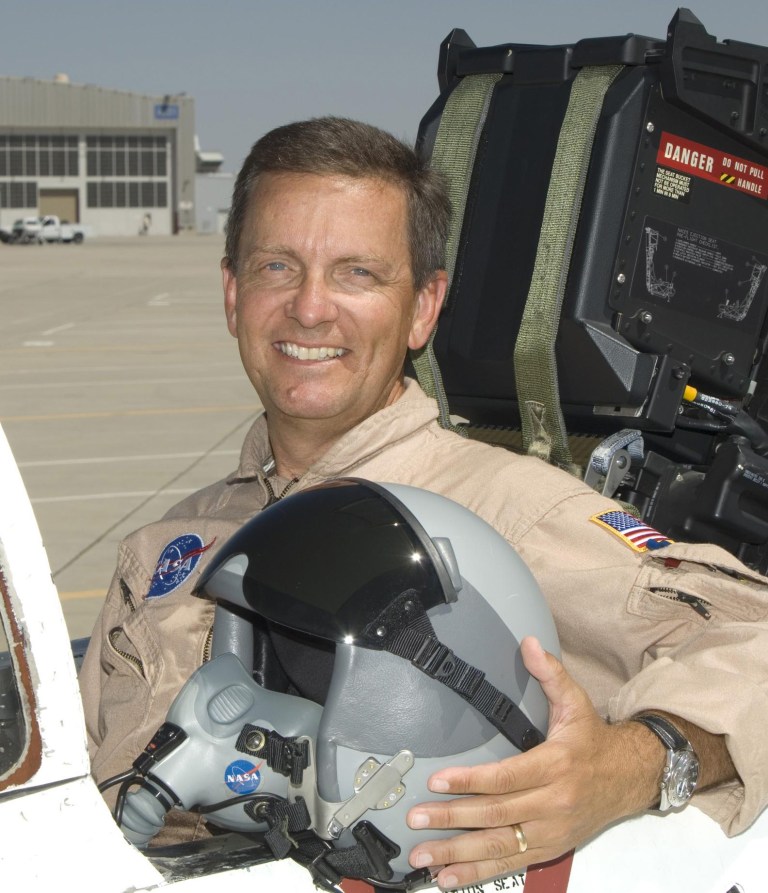 Man in flight suit holding helmet and posed in cockpit.