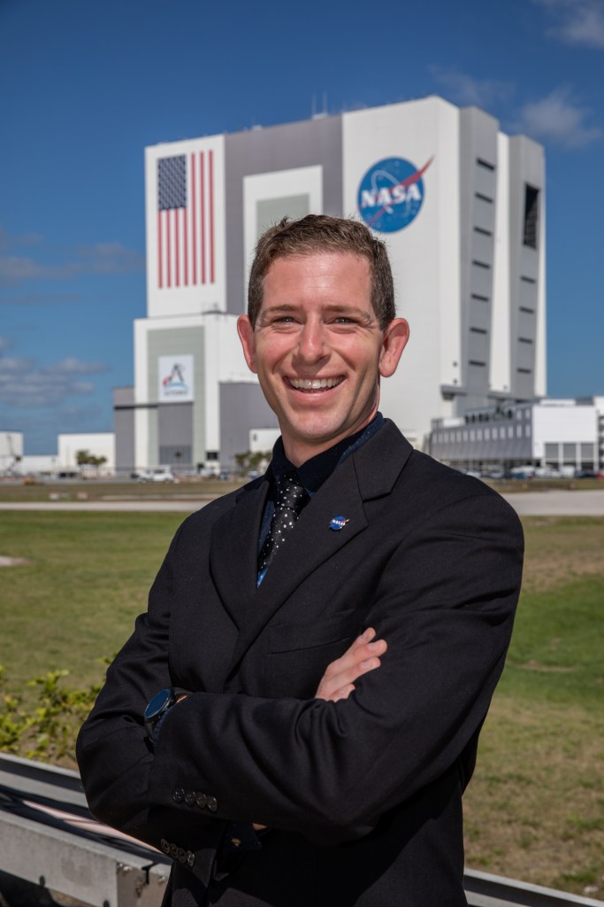 A photograph of Kennedy Space Center's Jesse Berdis with the Vehicle Assembly Building in the background.