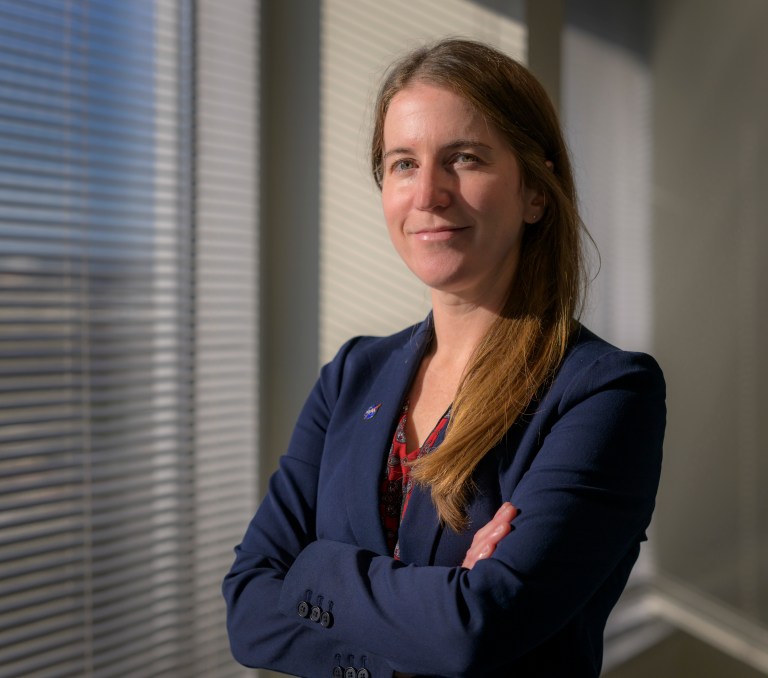 A smiling woman in a blue blazer stands with arms crossed next to an office window.