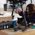 A researcher works to assemble a small spacecraft technology payload perched on a wooden tabletop in a ground-based technology facility.