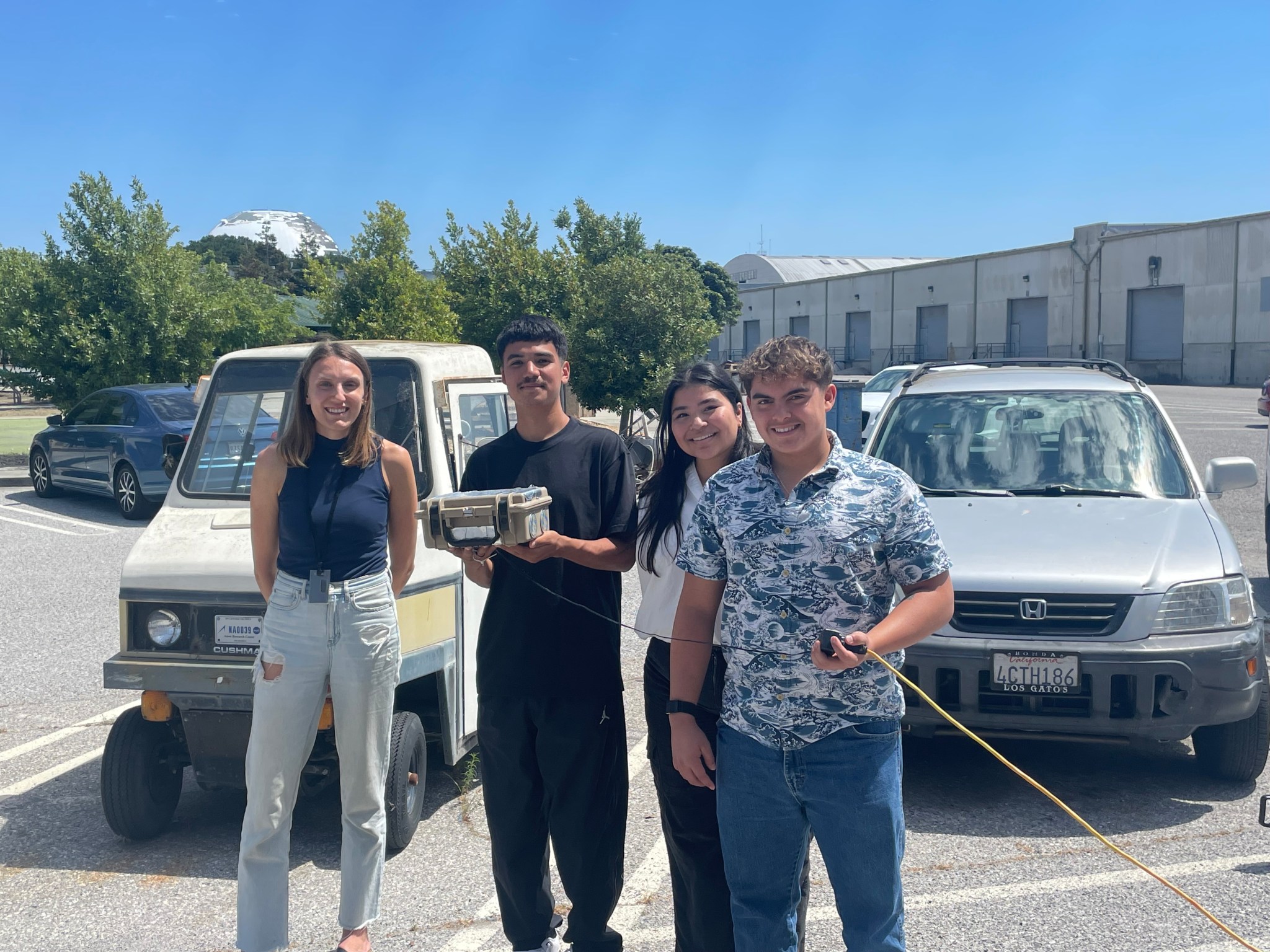 Four people stand in a parking lot, with two cars in the back. One in the middle, dressed in all black holds a tan and silver box about the size of a lunchbox.