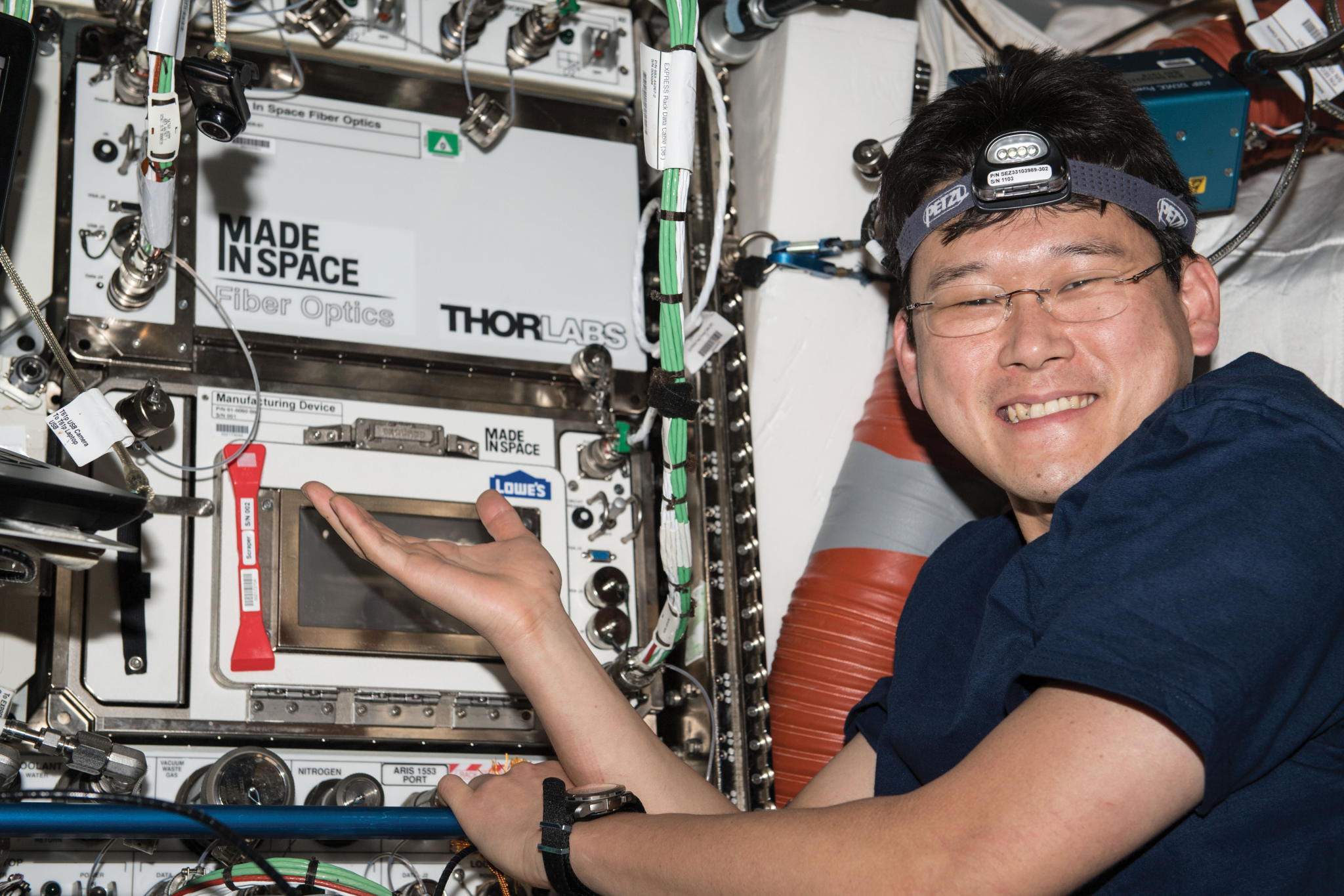 Kanai wears a dark blue short-sleeved t-shirt, a wristwatch, and a headlamp. He is smiling at the camera and gesturing toward hardware that has two microwave oven-sized sections, one above the other. The top one is white and is labeled “Made in Space Fiber Optics” and the other has a clear door and a large red handle. There are multiple cables and connectors around the hardware.