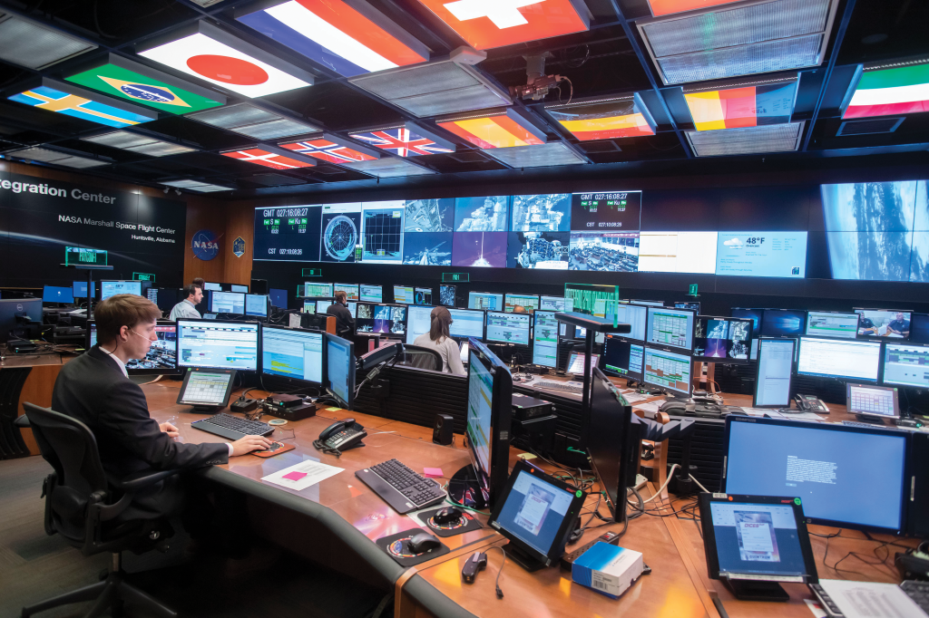 Team members sit in an open room filled with computer monitors and screens and flags from countries that participate in the ISS are on the ceiling.