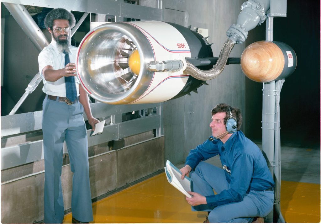 African American man stands next to aircraft engine inlet model inside a wind tunnel test chamber. Another man with headphones, kneels beside the model with notebook in hand.