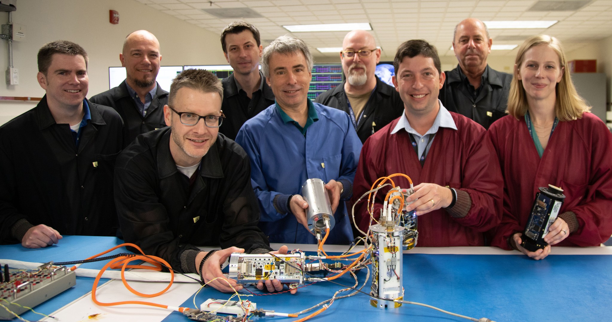 Group photo of nine smiling people, with some of those in the front row holding small, silvery cylinders (each a bit larger than a soda can) bespangled with circuitry; coils of wire spill out from some of the canisters.