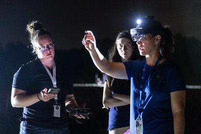 Three people in the dark. Two are observing the third person wearing a Augmented Reality headset and is pointing forward.