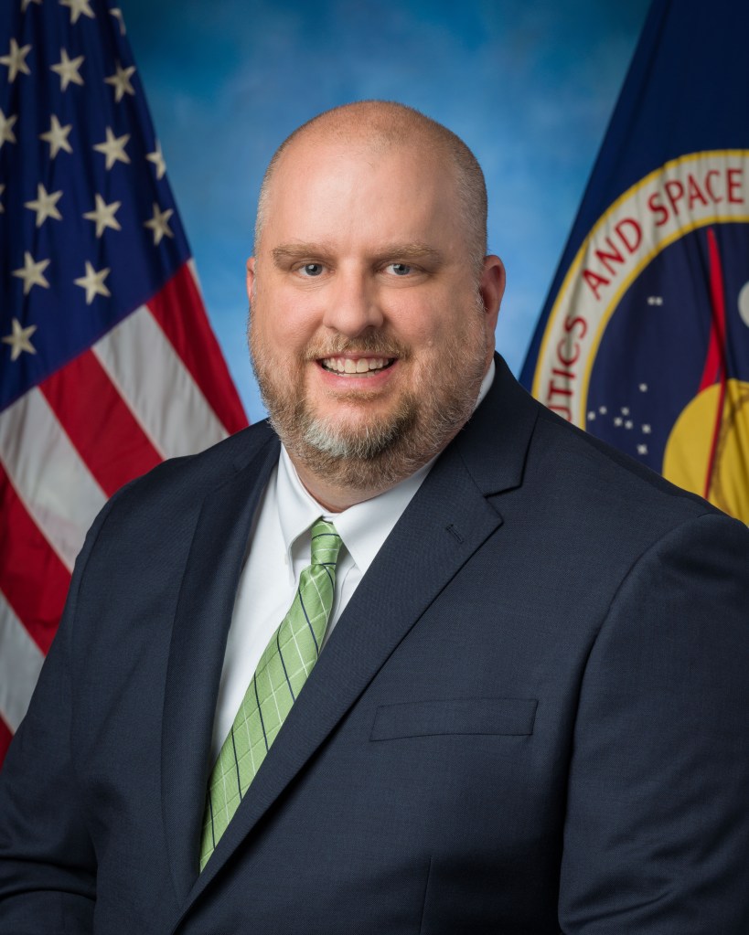 A bearded, bald man in a suit and tie poses in front of the American and NASA flags