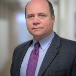 A white man with grey hair on the sides of his head. He's wearing a grey suit jacket, light blue shirt, and a solid purple tie.