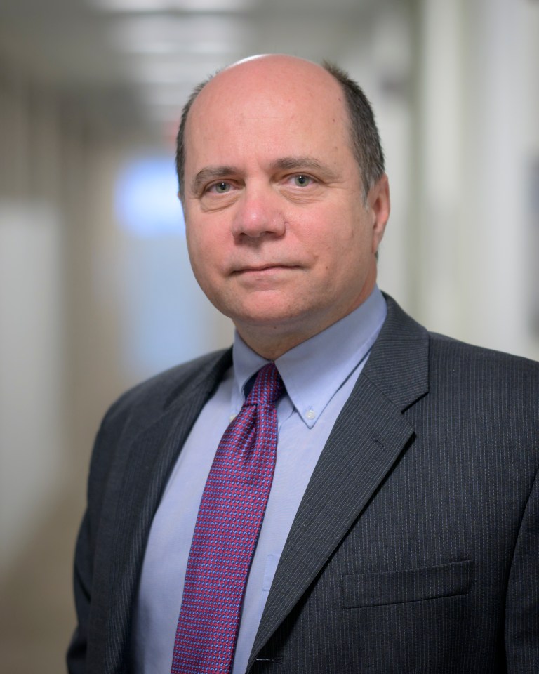 A white man with grey hair on the sides of his head. He's wearing a grey suit jacket, light blue shirt, and a solid purple tie.