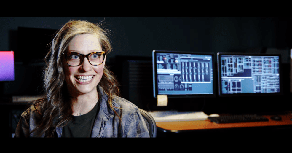 A young woman with glasses and long brown hair sits in front of computer screens smiling.