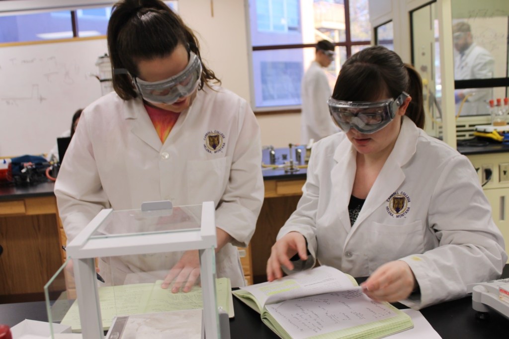 两名学生戴着护目镜，穿着印有大学标志的白色夹克，正在进行一个项目。
