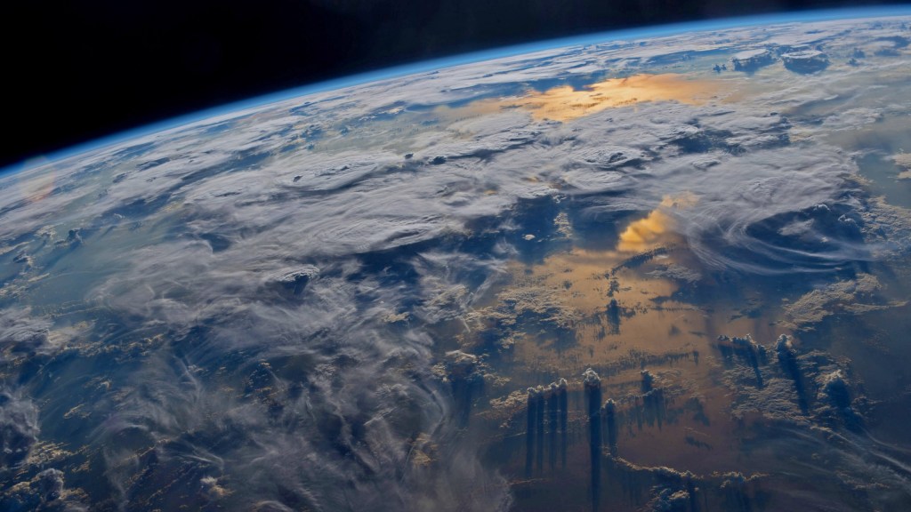 ISS photo of Earth's limb, showing clouds over the sea and sunset glint on the waters below.