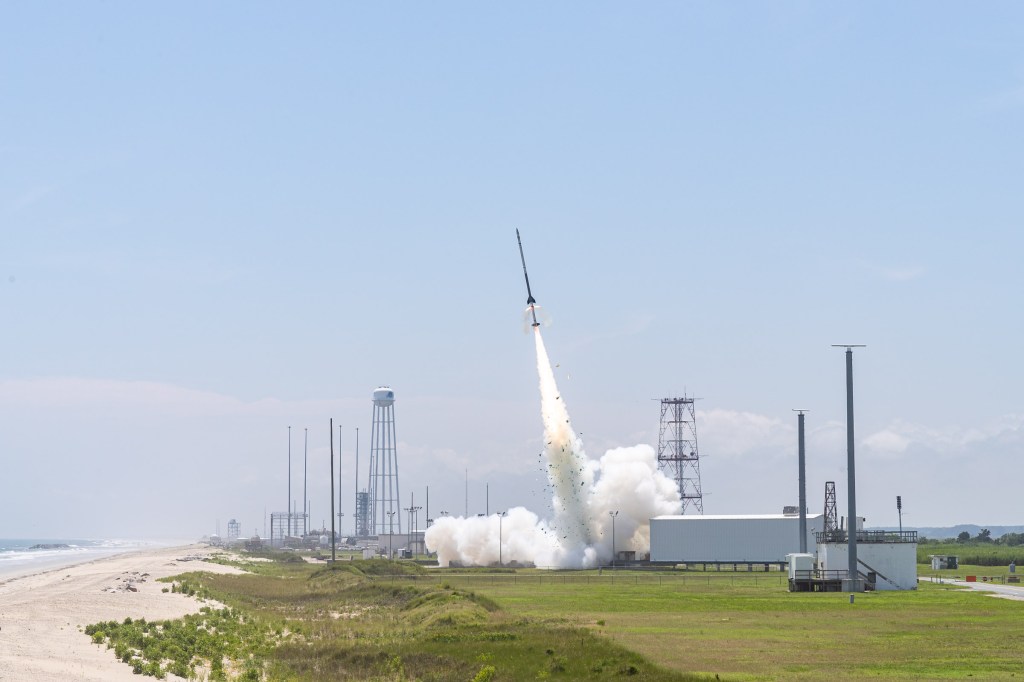 Landscape photo of a sounding rocket seconds in mid-launch off the launch pad with a bright white plume underneath. In the background is a clue blue sky. A beach line is seen on the left of the image, and various buildings and tall structures are farther in the background.