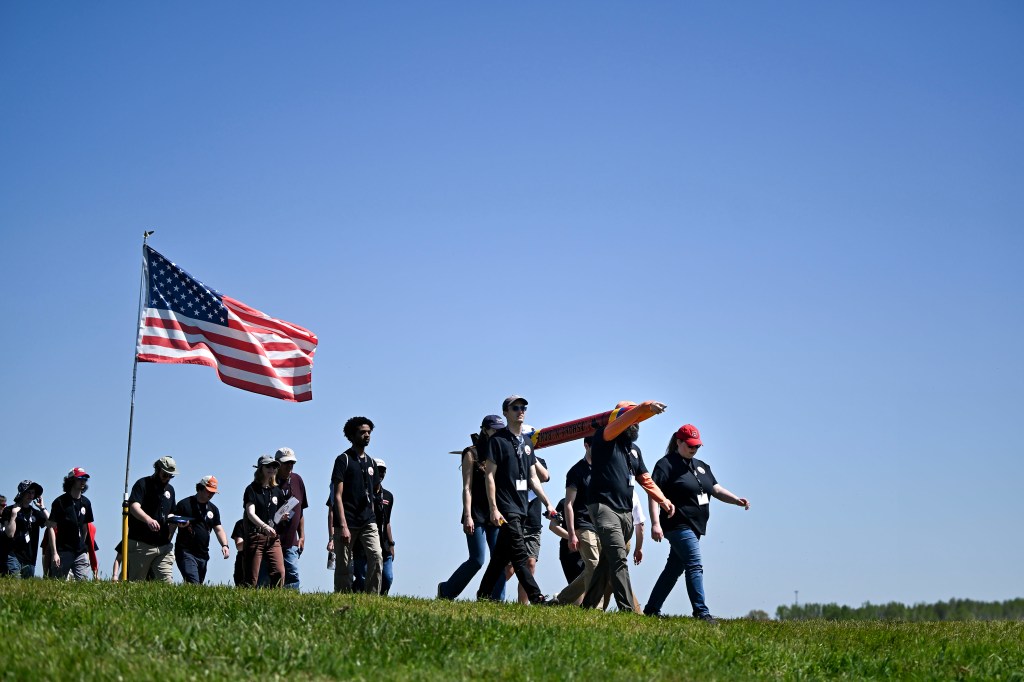 A group male and female students walk along a grassy field in Huntsville, Alabama with a Rocket as part of NASA's Student Launch. The American flag is in the foreground.