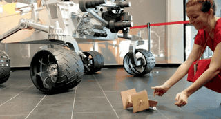 Image of a woman releasing a cardboard rover that jumps forward. A model of the Curiosity rover can be seen in the background