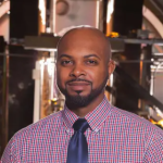 Man in shirt and tie looks at camera while standing inside a laboratory vacuum chamber.