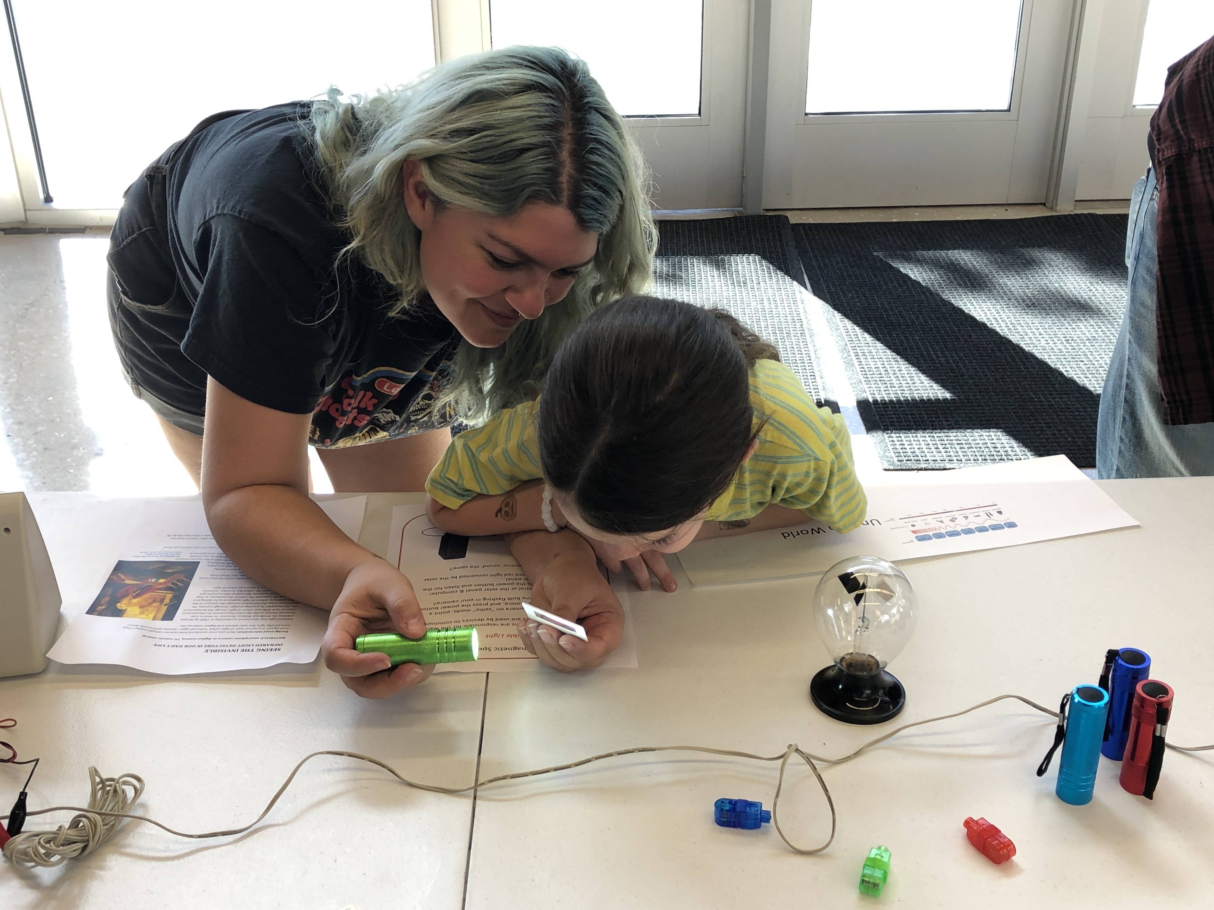 A parent and child lean in closely over a table to look at a flashlight <a href=