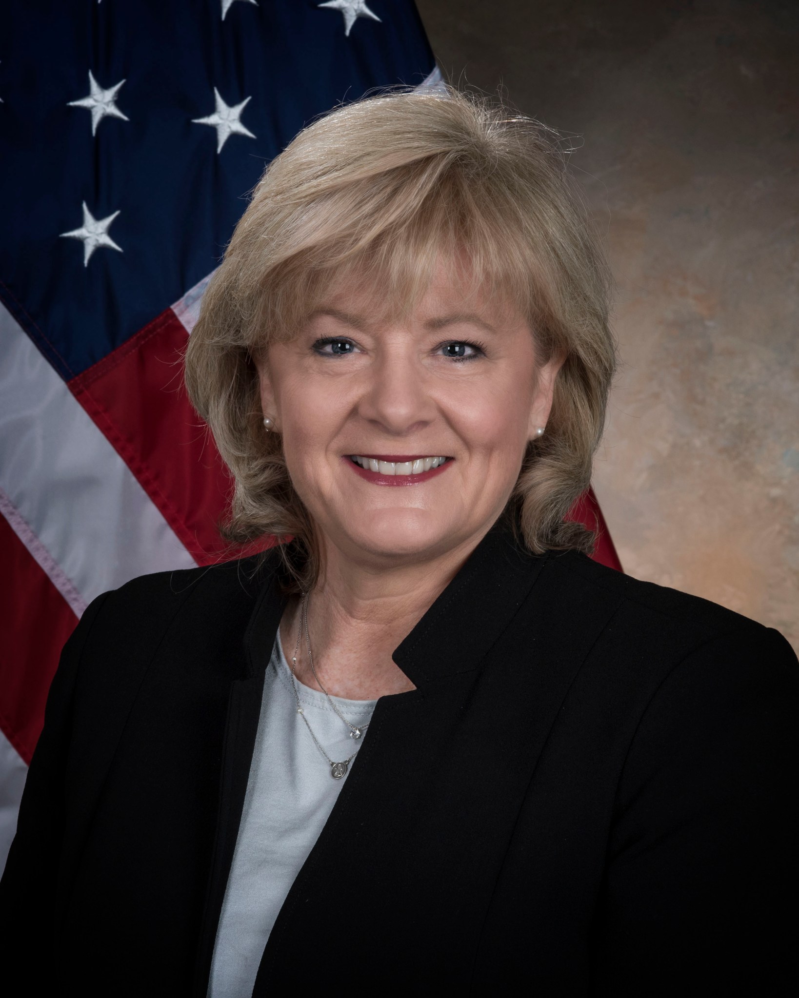 A photo of Jody Singer from Marshall Space Flight Center in front of an American Flag wtih a brown background behind her.