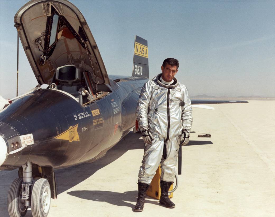 A man wearing a silver space suit stand next to the X-15  aircraft.