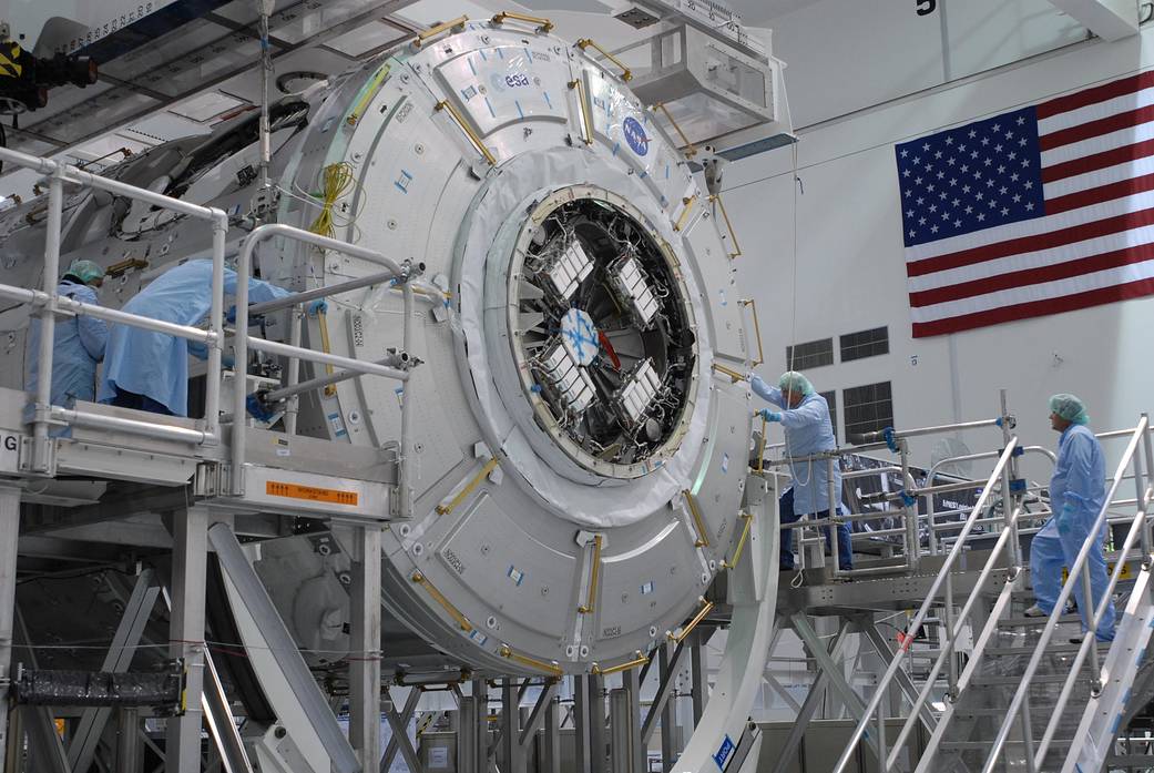 In the Space Station Processing Facility at Kennedy Space Center, an overhead crane lowers the Tranquility module, or Node 3, toward a work stand. 