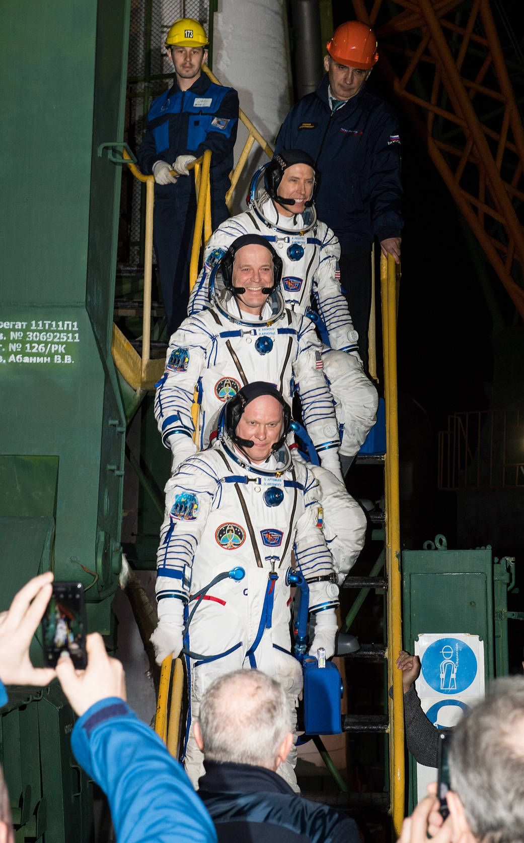 Three crew members in Sokol suits ascend ladder and wave at crowd