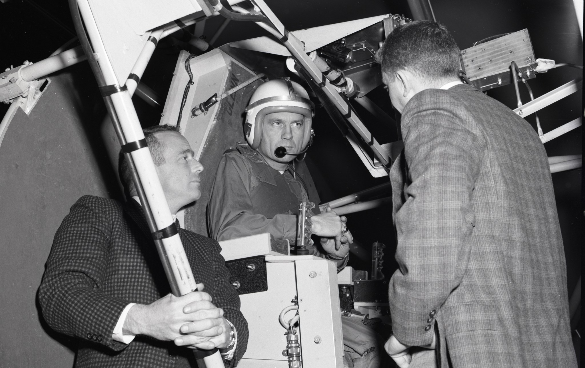 Man with helmet in pilot chair talking with two men in suits.