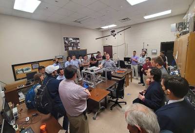 A group of university students and faculty gather around a laboratory workbench.