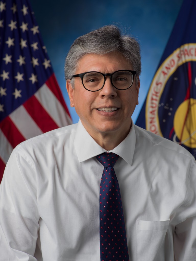 A portrait of a man wearing glasses, a white dress shirt, and a navy tie with red dots. He is seated in front of the U.S. flag and NASA’s official emblem.