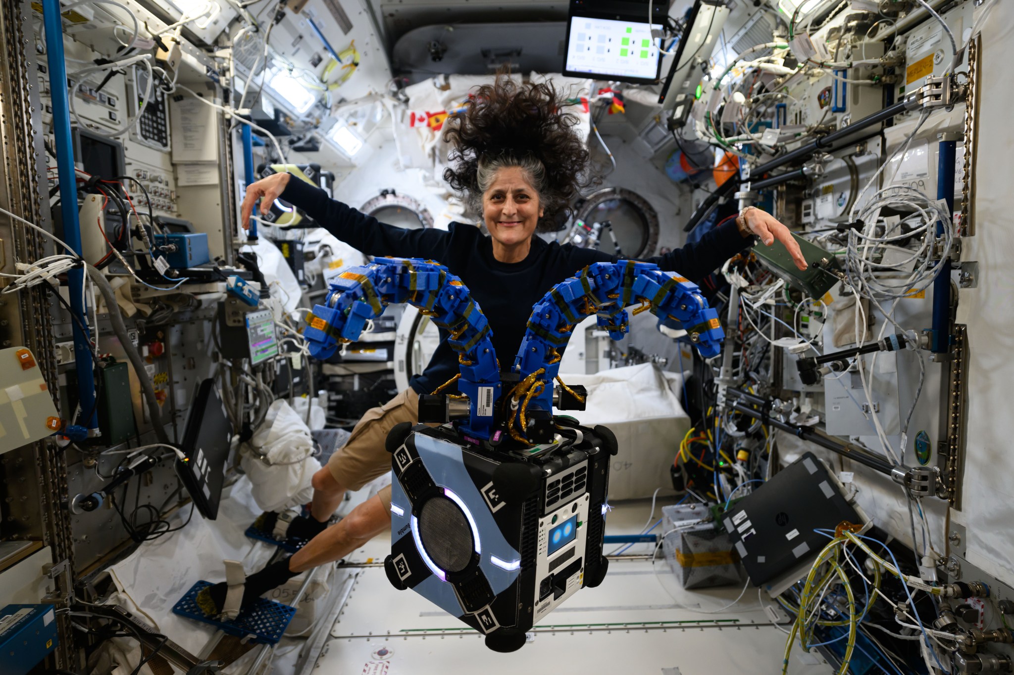 An Astrobee robot, a blue and black cube the size of a small carry-on, floats in the front of this image. It has two multi-jointed blue arms extending from its top like tentacles. Williams, smiling at the camera and wearing a long-sleeved black shirt and khaki shorts, floats behind the robot and holds her arms out in imitation.