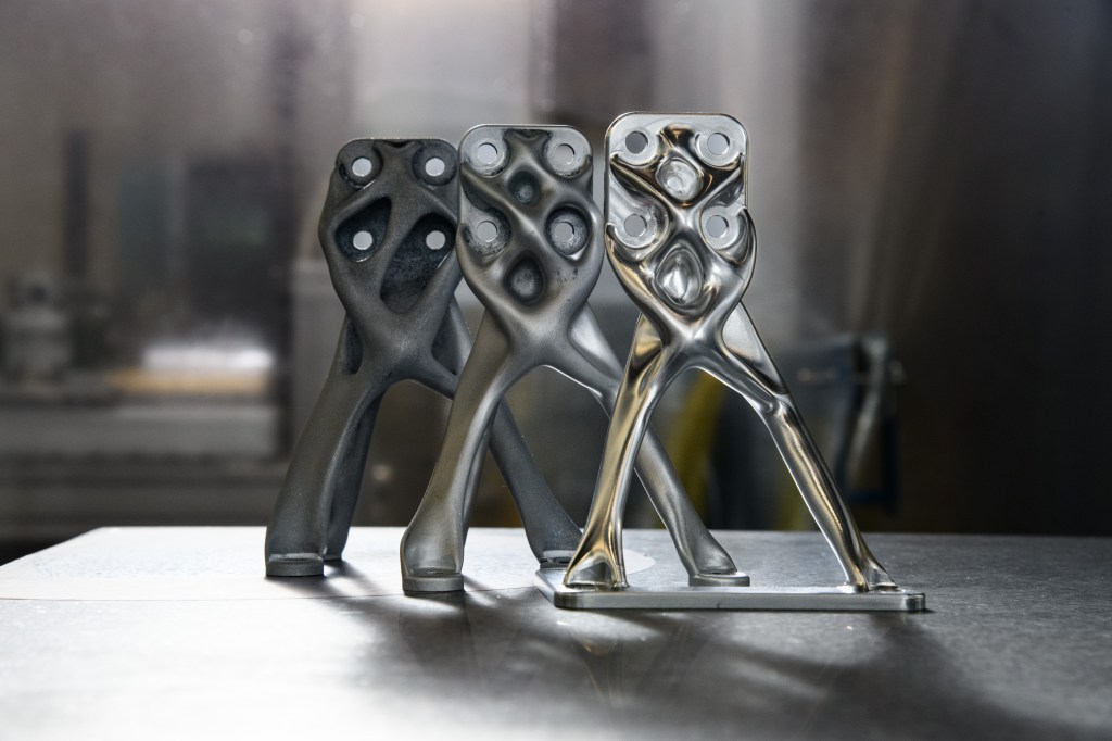 Three organic-looking structures of gray metal stand on a smooth table with a blurred, laboratory background. The pieces look identical, but they range from shiny silver metal in the front to matte gray metal in the back.