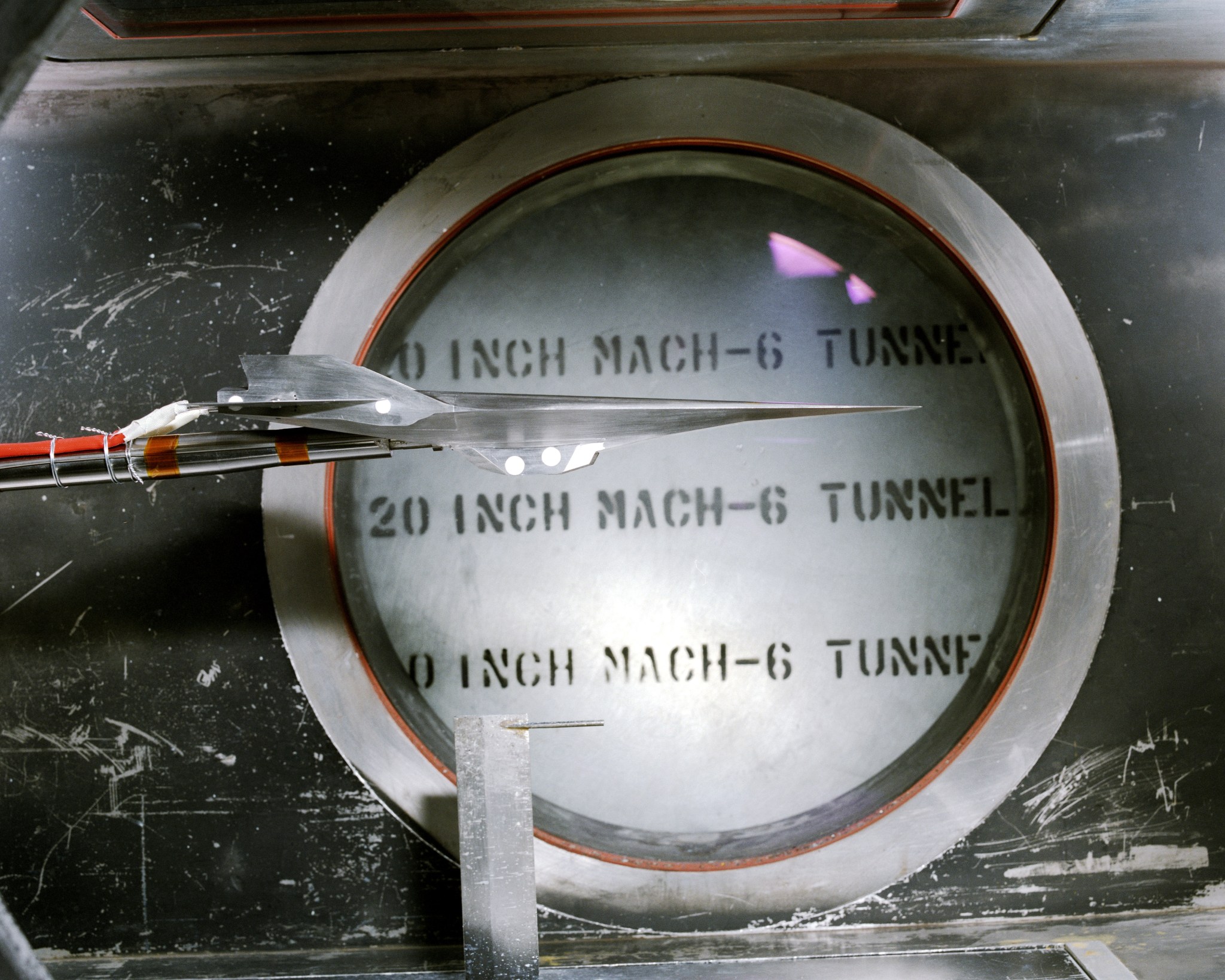 A steel model of a hypersonic vehicle and sensor in front of a window in a wind tunnel labeled the 20 inch Mach 6 Tunnel. The model is narrow and sharp.
