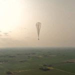 Aerostar’s high-altitude balloon is shown floating above expansive green fields and a small body of water appears in the lower left corner. The sun, low in the sky and out of frame, is casting sunlight through clouds and haze.