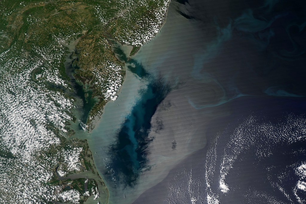 Satellite image of the Delmarva Peninsula area of the East Coast of the U.S., which is a green and brown coastline with the Delaware and Chesapeake Bays. Most of the land is covered in small puffs of clouds. The majority of the image is blue ocean water in the Atlantic. There is a large, gleaming patch of silvery water that looks almost like a mirror, labeled "sunglint." Next to it is an exceptionally colorful patch of ocean water, including teal swirls of phytoplankton, where there is no sunglint, which are labeled "phytoplankton." A darker green and blue patch next to the sunglint is labeled "low wind."