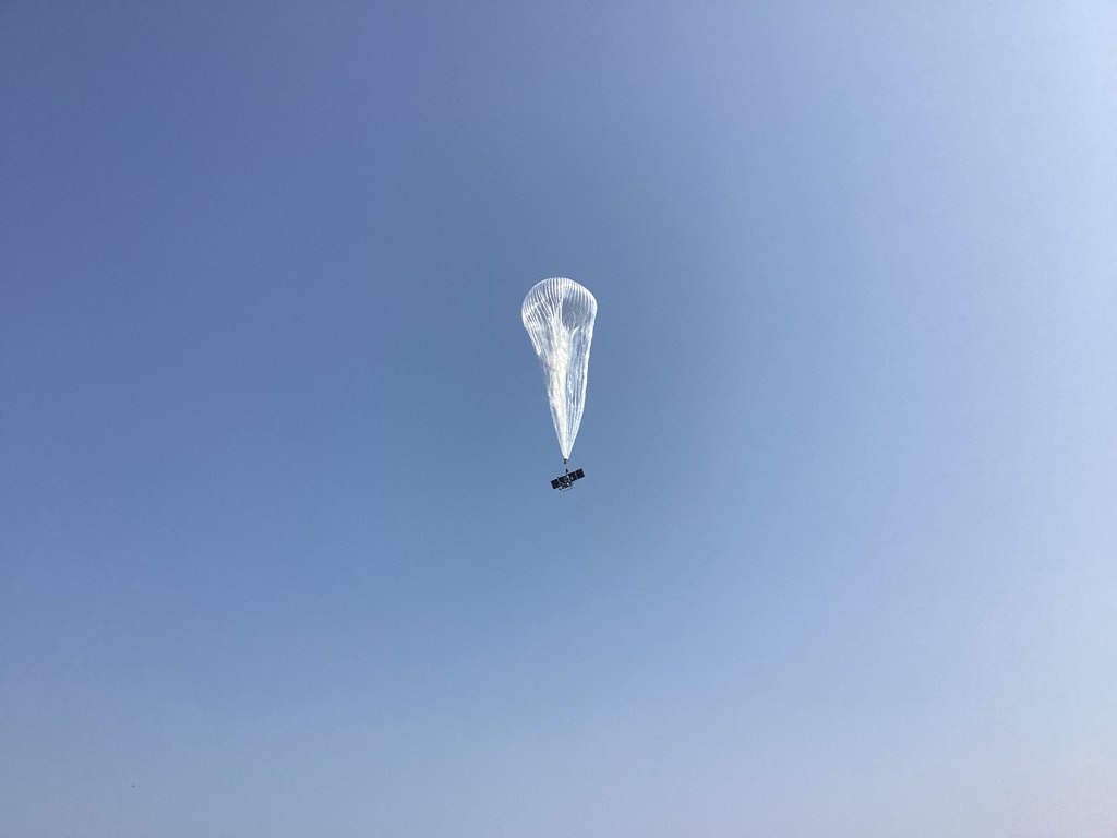 A silvery, translucent balloon with the payload gondola visible beneath it rises into the sky.