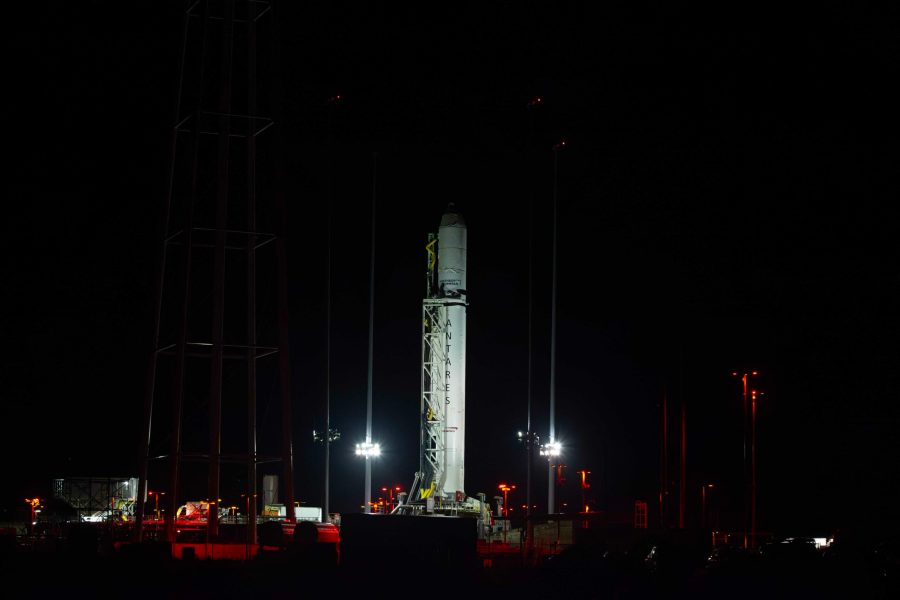 The Antares rocket stands on its launch pad at night. The rocket is white and the name "Antares" is printed vertically in all caps down the side.