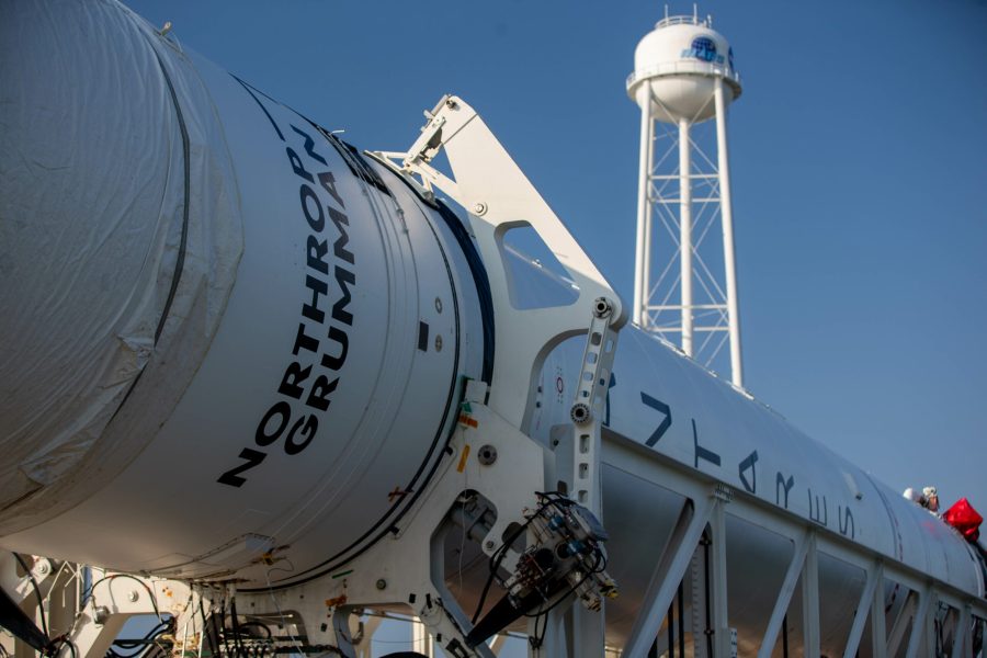 Close up of the Antares rocket on its side with a white water tower in the background. The rocket is all white with Northrop Grumman's logo and Antares written vertically down one side. Behind the the sky is a light blue.