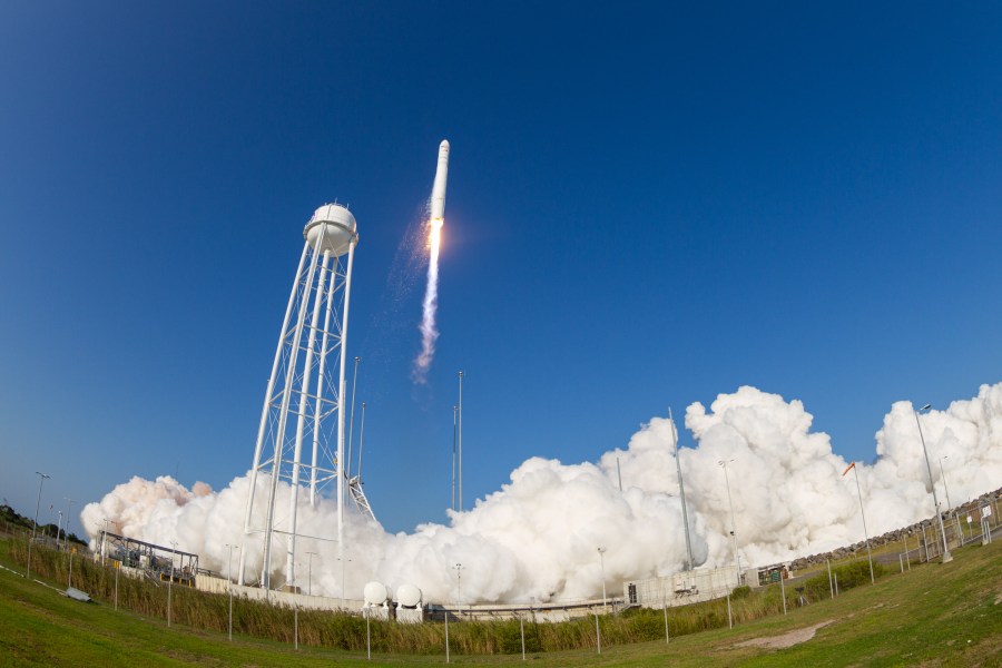 A large, white rocket lifts off from a launch pad against a bright blue sky. White flame and exhaust streams out from its base with billowing clouds of exhaust on the launch pad. A tall water tower stands next to the launch pad. In the foreground, tall green grasses behind a fence line.