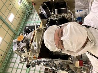 A man with glasses, wearing white hooded coveralls and a white mask, takes a selfie in front of a spacecraft in a clean room. The spacecraft is gold and sits on a tall table. The walls are gridded with filter panels.