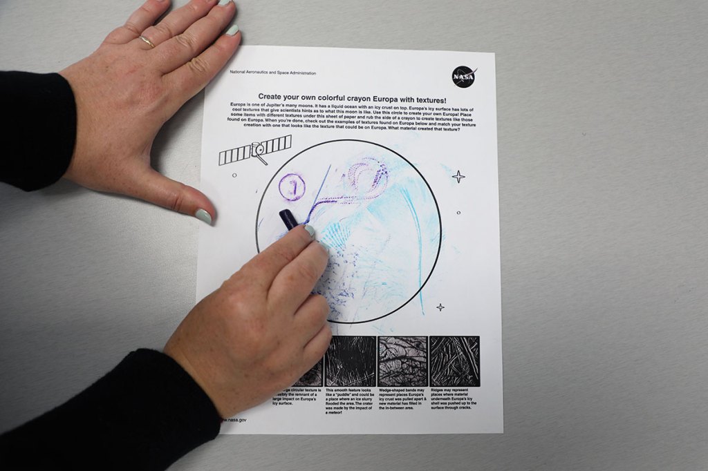 A person uses the side of a crayon to create a textured rubbing on a “Create your own colorful crayon Europa with textures!” worksheet. The resulting textured collage represents the rough icy surface of Jupiter’s moon, Europa