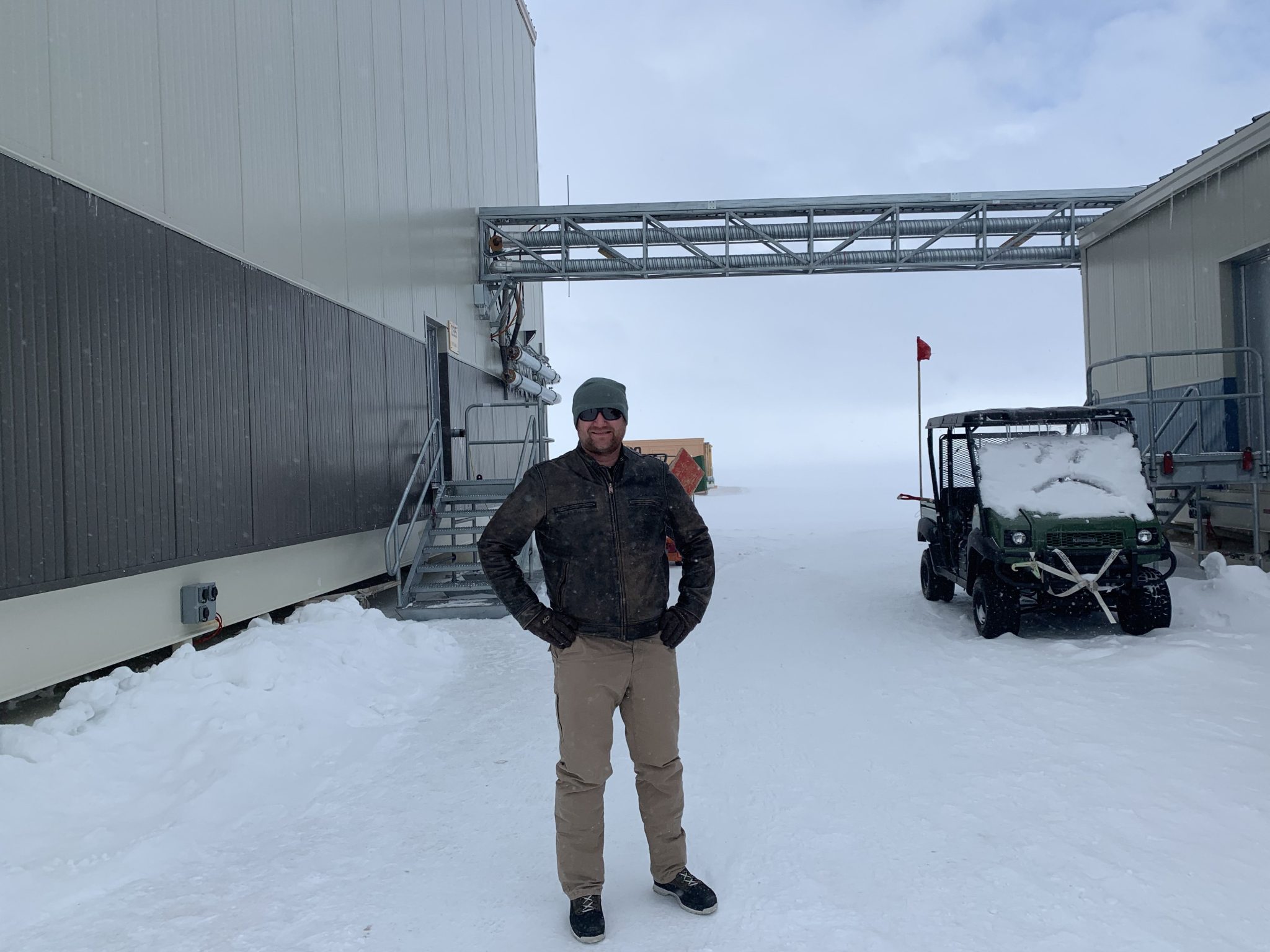 A man in winter clothes stands between two buildings and smiles, with his hands in his pockets. The ground is covered in snow, a small golf cart behind him is covered in snow, and the horizon is obscured by blowing snow.