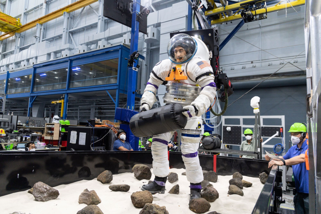 A volunteer from NASA’s Artemis Extravehicular Activity training group moves a 30-pound object through a boulder field while in a spacesuit connected to NASA’s Active Response Gravity Offload System, or ARGOS.