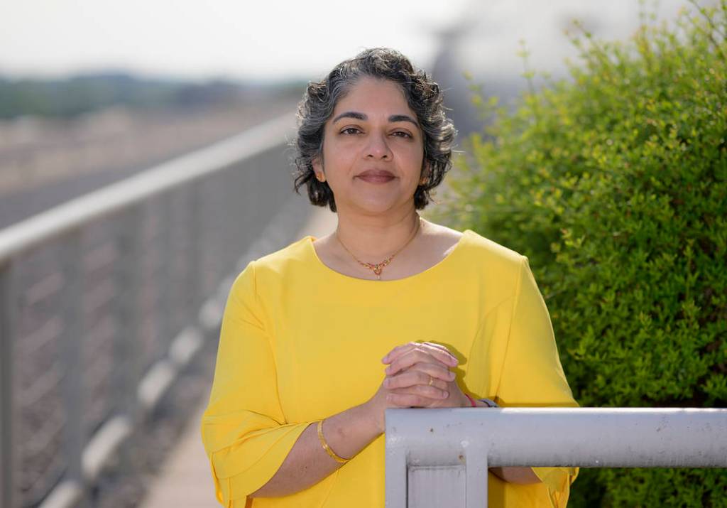 An Indian woman with short, curly, salt and pepper hair smiles softly at the camera wearing a sunshine yellow quarter sleeve blouse. Her hands are clasped as she leans agains a railing outside, with bright green bushes on her right-hand side.